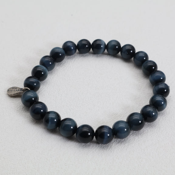 Top view of the Midnight Focus blue tiger’s eye bracelet with glossy blue-black beads and a silver charm on a white surface.