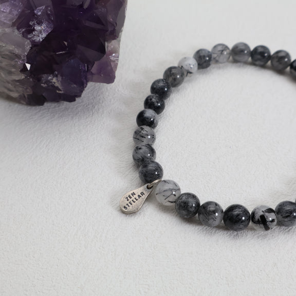 Close-up of black needle-like inclusions in quartz beads and a ZenStellar silver charm beside a purple crystal cluster.