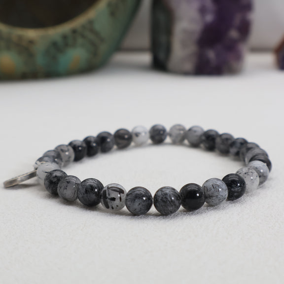 Shadow Veil black rutilated quartz bracelet with translucent gray beads, black inclusions and a silver charm, shown from a low angle.
