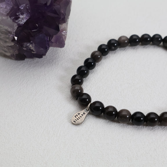 Close-up of polished black obsidian beads and a ZenStellar silver charm beside a purple crystal cluster.