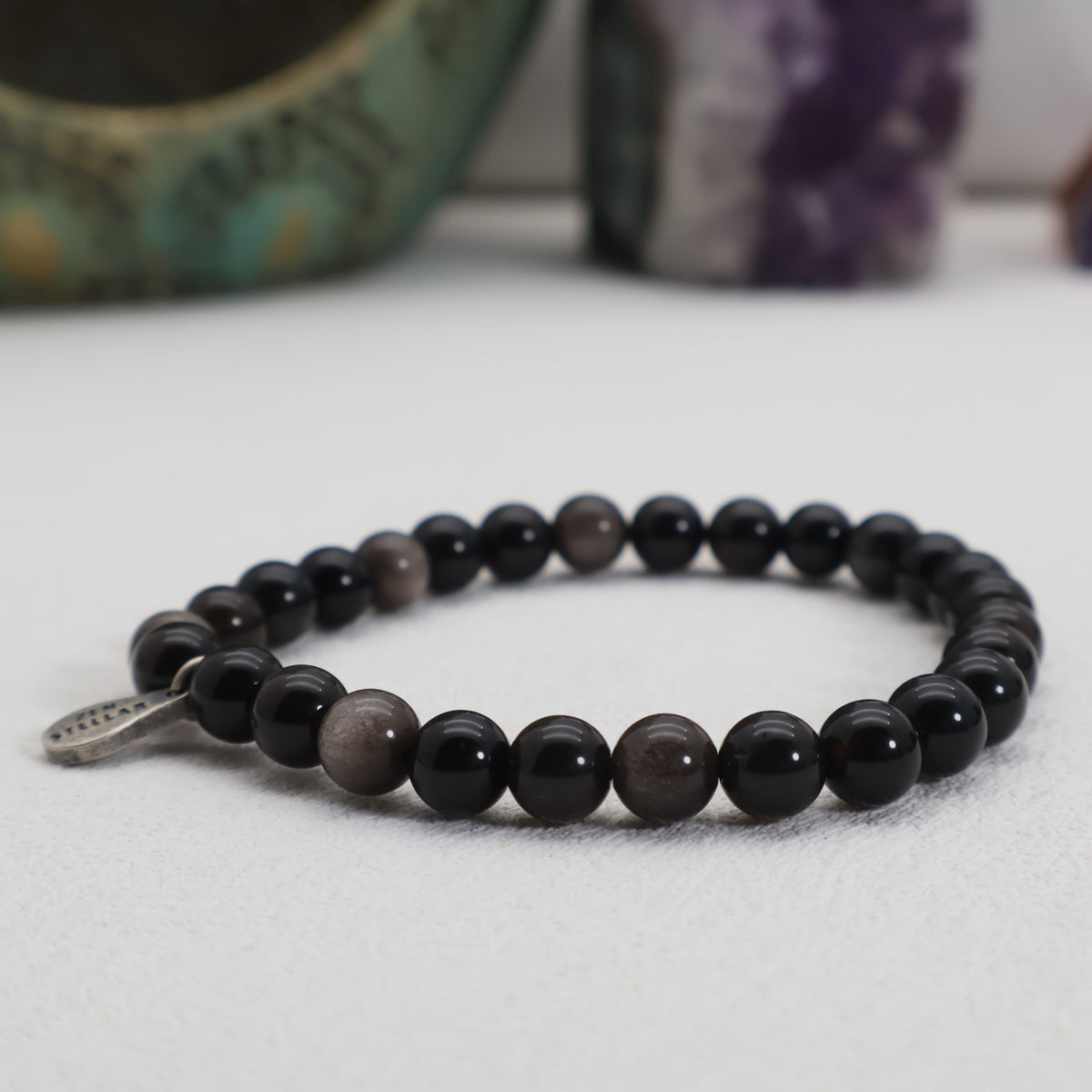 Silver Shadow obsidian bracelet with polished black beads, subtle gray sheen and a silver charm, shown from a low angle.
